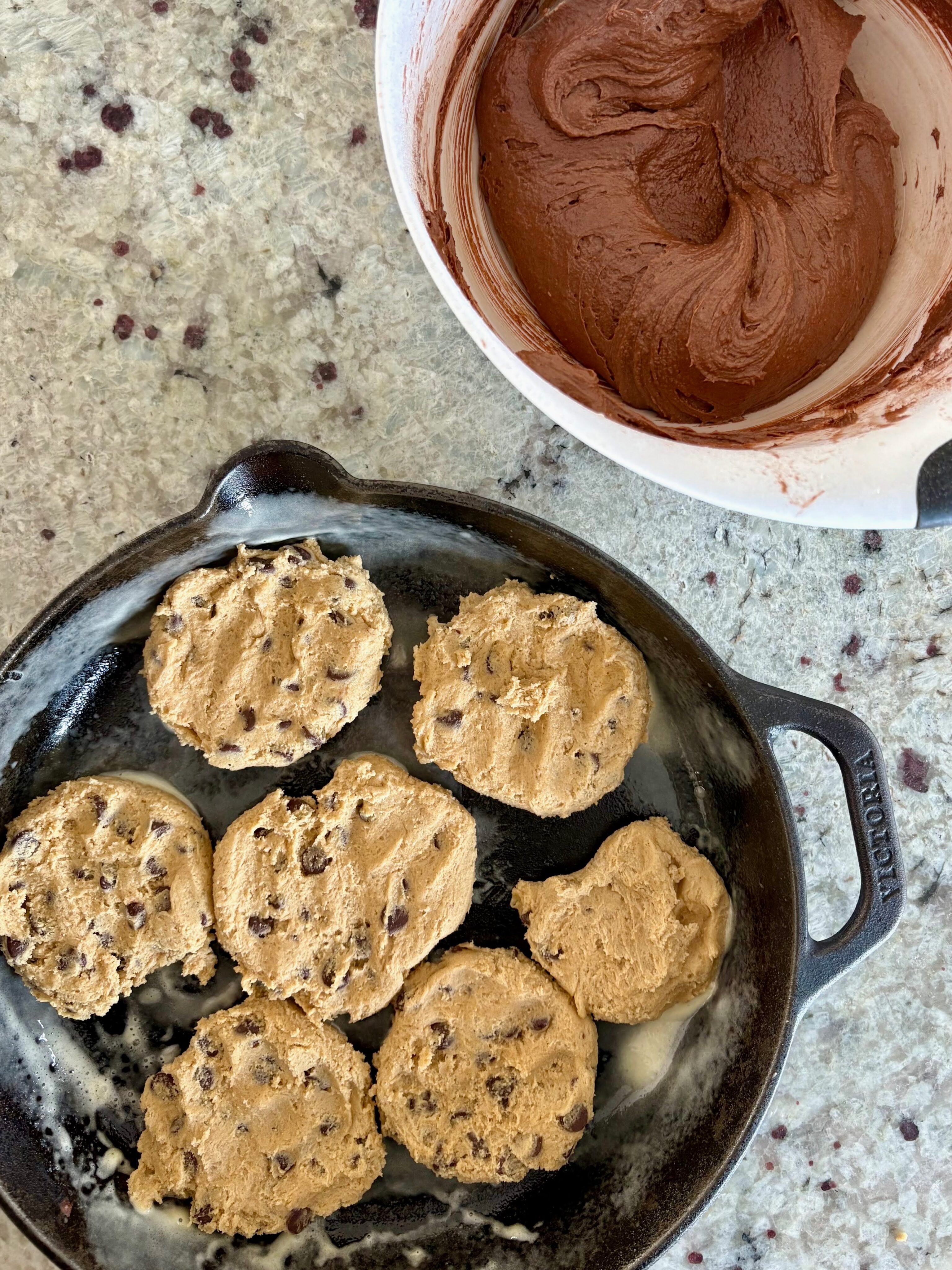 Process shot of the skillet brookie. The chocolate chip cookie dough has been pressed into the skillet. The brownie batter is set aside in a bowl next to the skillet.